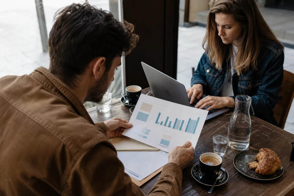 Two co-founders reviewing a printed monthly revenue report on a cafe table